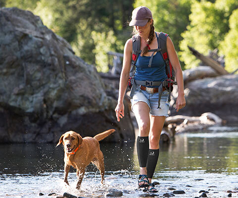 A woman with calf sleeves walks her dog through shallow water