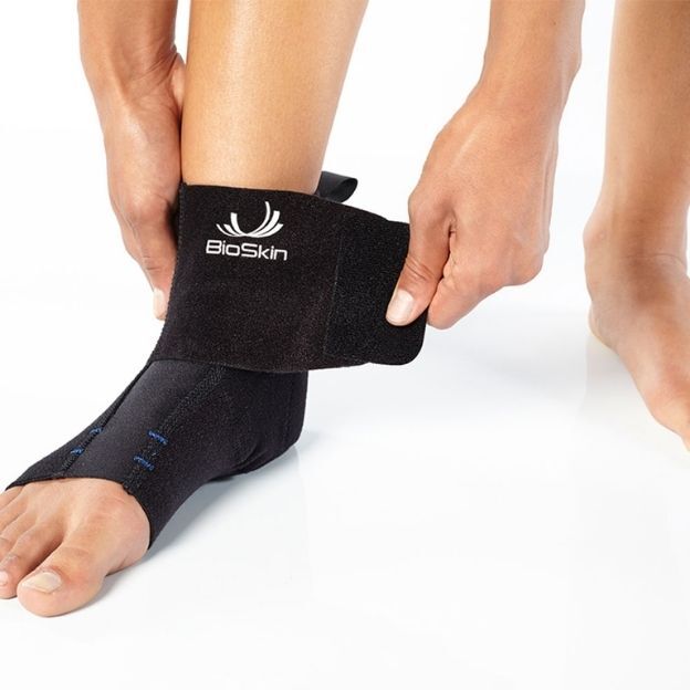 Close-up of person applying a black BioSkin ankle support, pulling the strap across the ankle for compression.