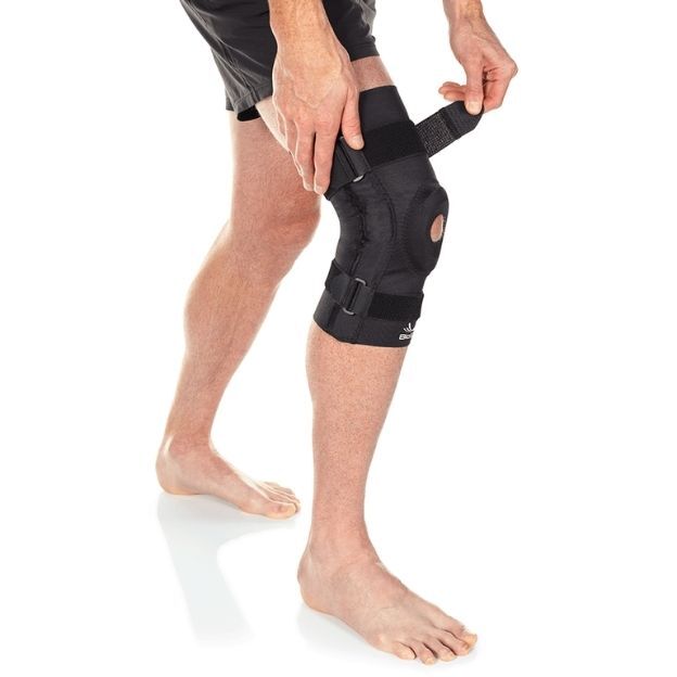 Barefoot person adjusts black hinged knee brace with open patella, tightening the top strap while standing.